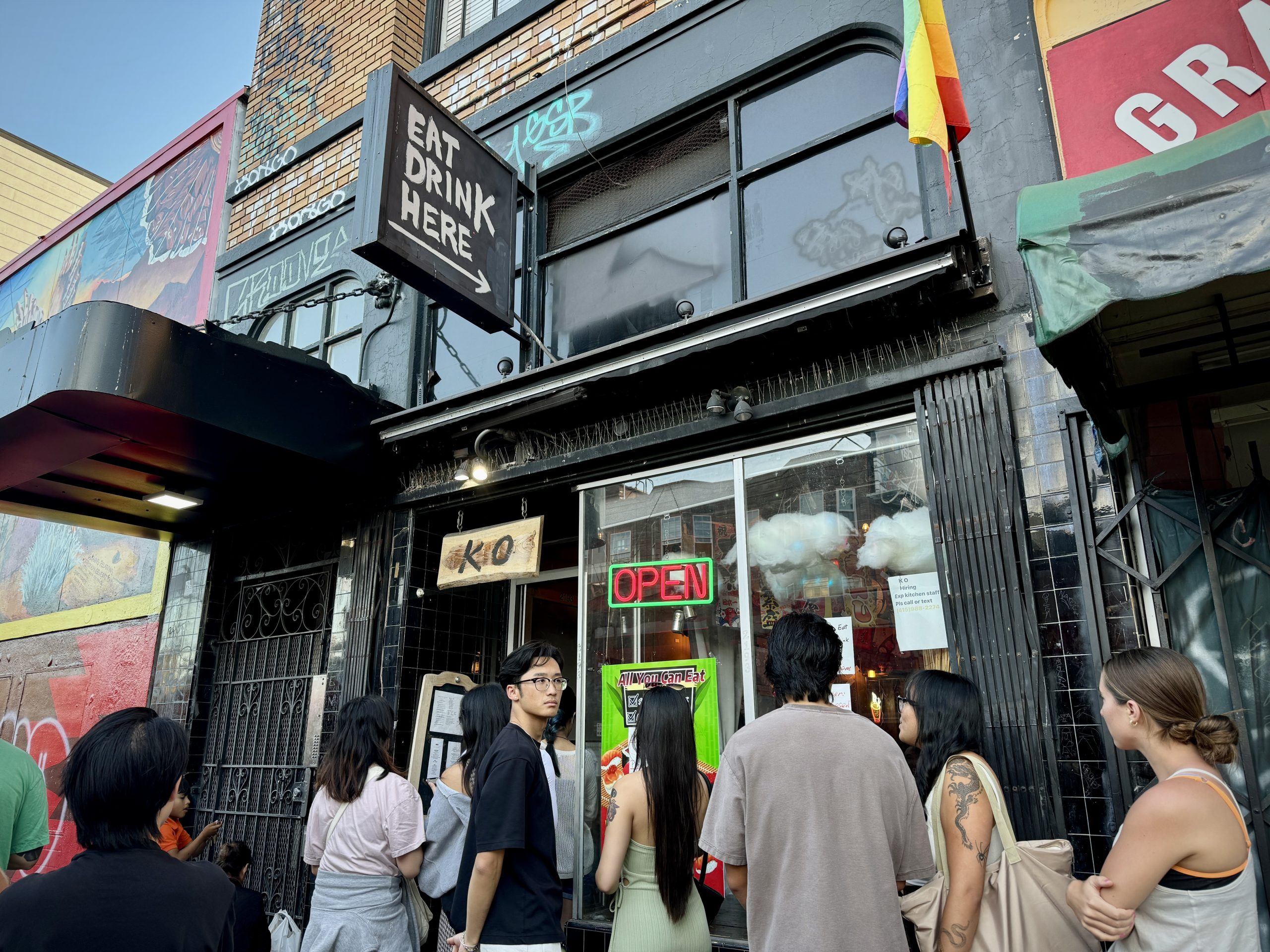 People waiting in line outside Ko, a restaurant with a sign reading "Eat Drink Here." The establishment has an "Open" sign and a small pride flag.
