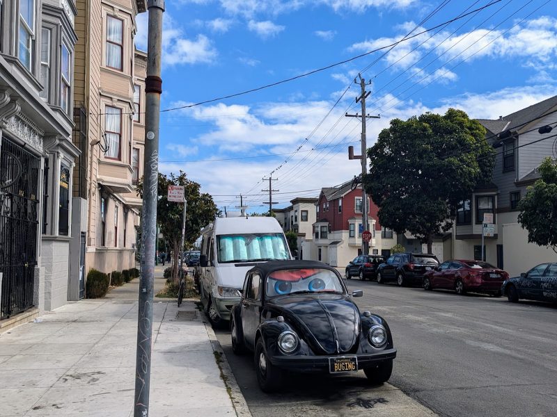 Street view with cars parked alongside the curb. A sign reads "End LUCKY." A classic black car is parked on the side under the sign. Blue sky with scattered clouds above.