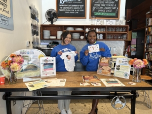 Two people in matching blue shirts stand behind a table displaying pamphlets, a baby onesie, and flowers. They're smiling indoors, with shelves and a menu board in the background.