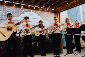 A mariachi band with six members plays string instruments under a wooden pergola. They are dressed in white shirts with red ties.