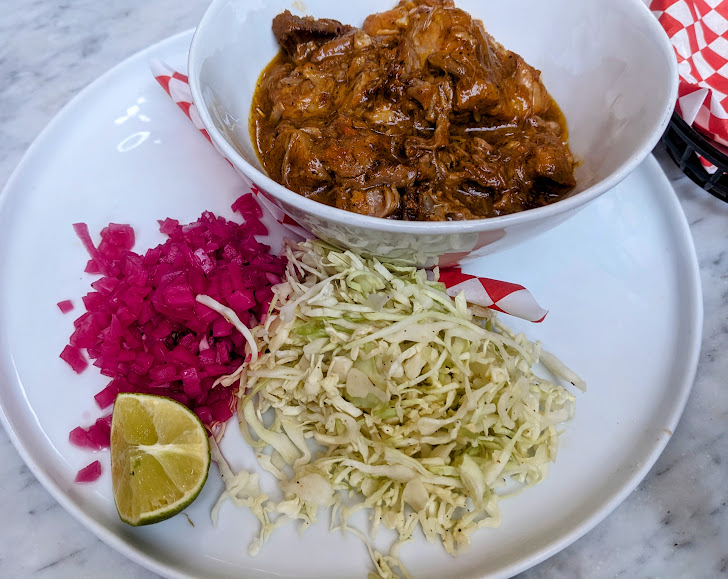 A bowl of shredded meat stew is on a plate with chopped red pickles, shredded cabbage, and a lime wedge.