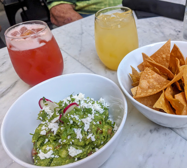 Bowl of guacamole topped with cheese and radish slices, next to a bowl of tortilla chips. Behind them are two drinks, one red and one yellow, on a marble surface.