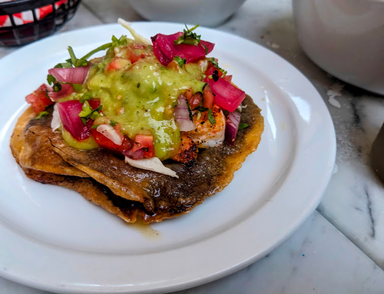 A plate of tacos topped with diced tomatoes, red onions, chopped herbs, and green sauce, on a white table.
