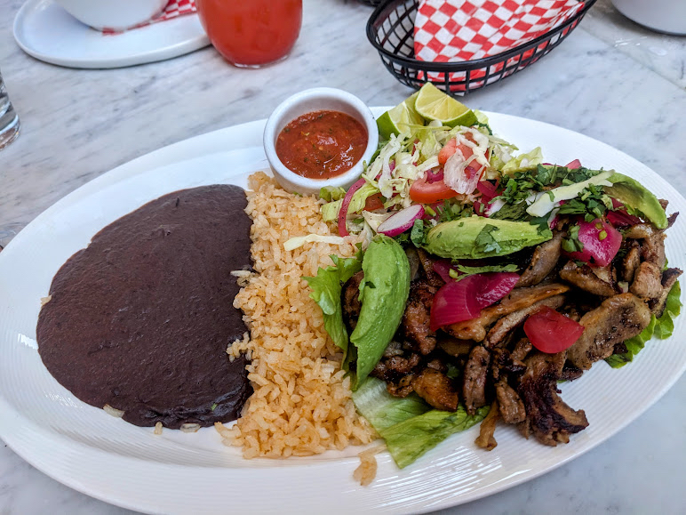 A plate of grilled meat with sliced avocado, pickled onions, and fresh greens, served with rice, refried beans, and a small bowl of salsa on the side.