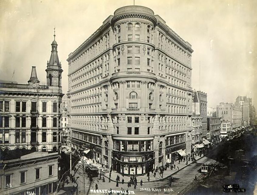 Historic photo of the James Flood Building at Market and Powell Streets, a large, multi-story structure with ornate details, surrounded by bustling city life and streetcars.