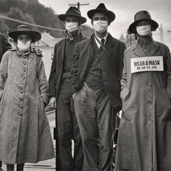 A group of four people standing outdoors, all wearing face masks. The person on the right has a sign on their coat that reads, "Wear A Mask Or Go To Jail.