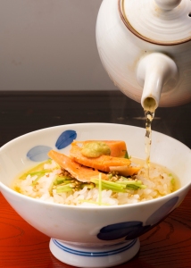 A teapot pours broth over rice topped with salmon and vegetables in a bowl.