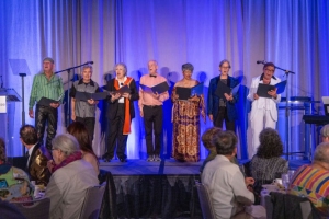 Seven people stand on a stage holding booklets in front of an audience, with a blue curtain backdrop.
