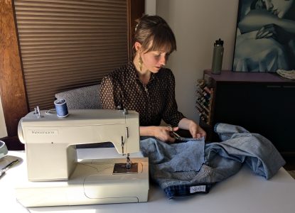 Audrey Danser of Salvage Studio sits at a table with a sewing machine, cutting fabric a denim patch for a pair of jeans.