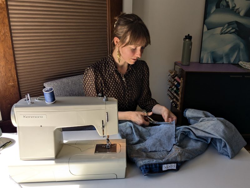 Audrey Danser of Salvage Studio sits at a table with a sewing machine, cutting fabric a denim patch for a pair of jeans.