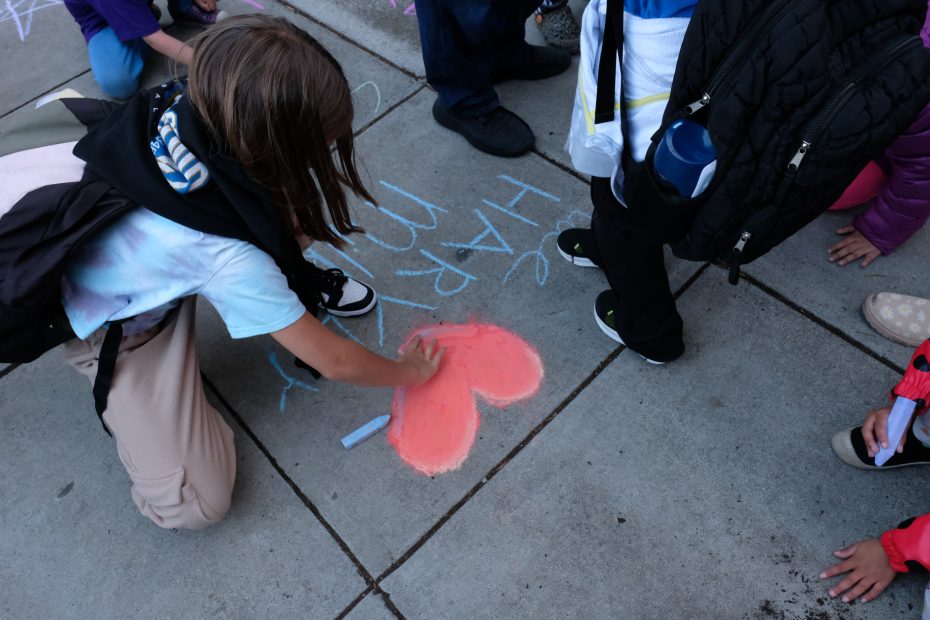 A child from SFUSD uses chalk to color a heart on the sidewalk, surrounded by several other children.
