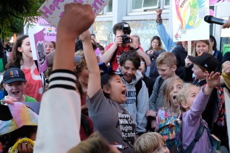 A group of enthusiastic children cheer, holding colorful signs celebrating Harvey Milk, while a photographer snaps pictures in the vibrant, crowded outdoor setting.