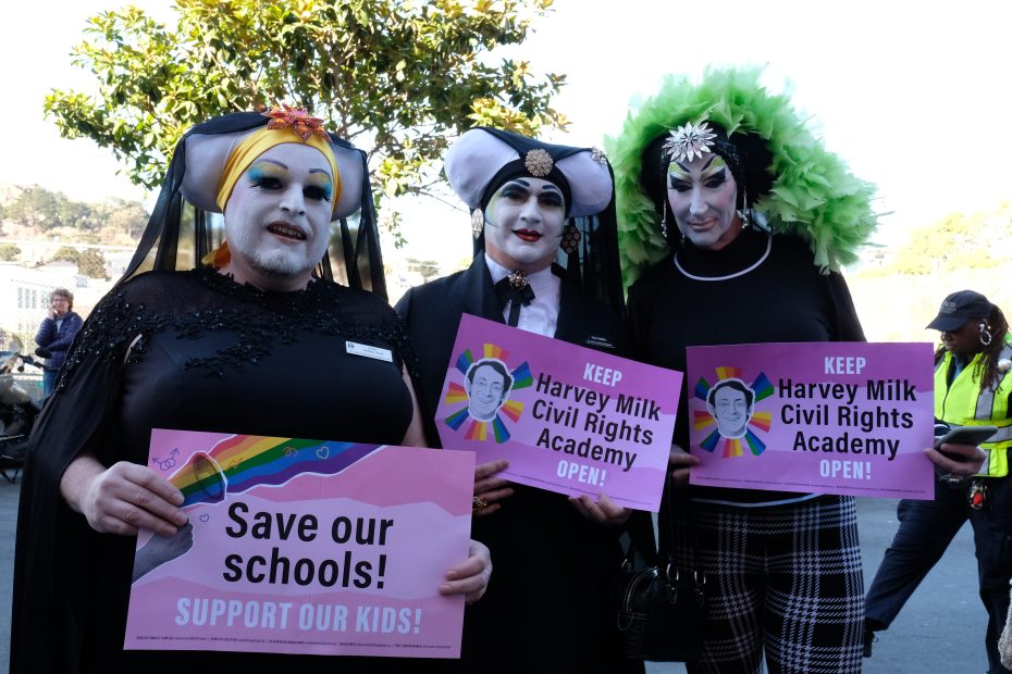 Three people dressed in colorful costumes hold signs supporting Harvey Milk Civil Rights Academy and advocating for school support.