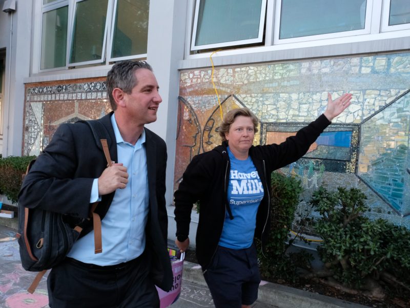 Two people stroll past a building adorned with mosaic art. One, in a suit and carrying a bag, appears ready for business. The other, dressed casually, gestures expressively as if sharing insights about sfusd.