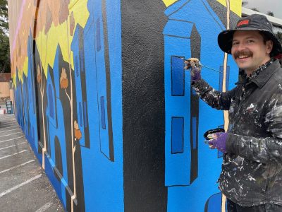 Person painting a colorful mural on a building wall, wearing a black hat and paint-stained clothing.