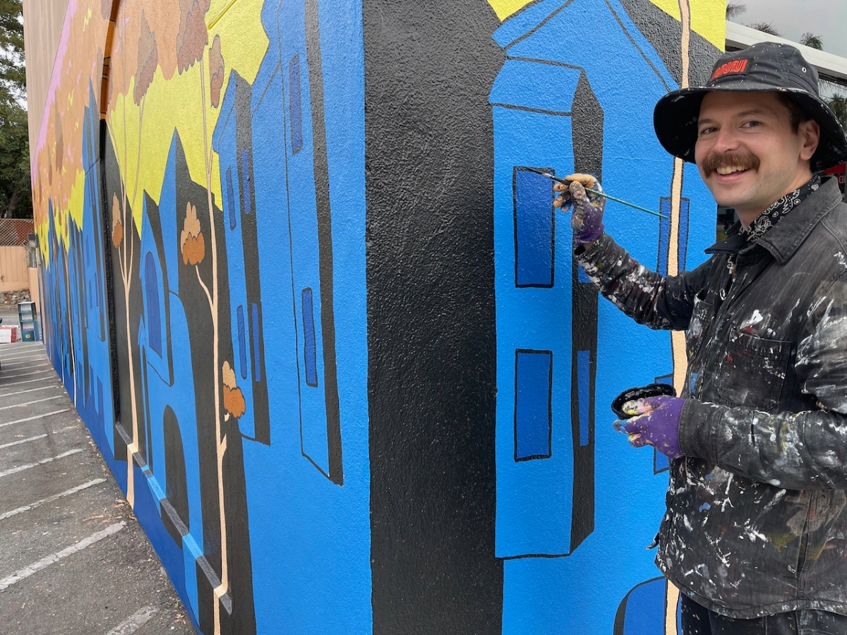 Person painting a colorful mural on a building wall, wearing a black hat and paint-stained clothing.