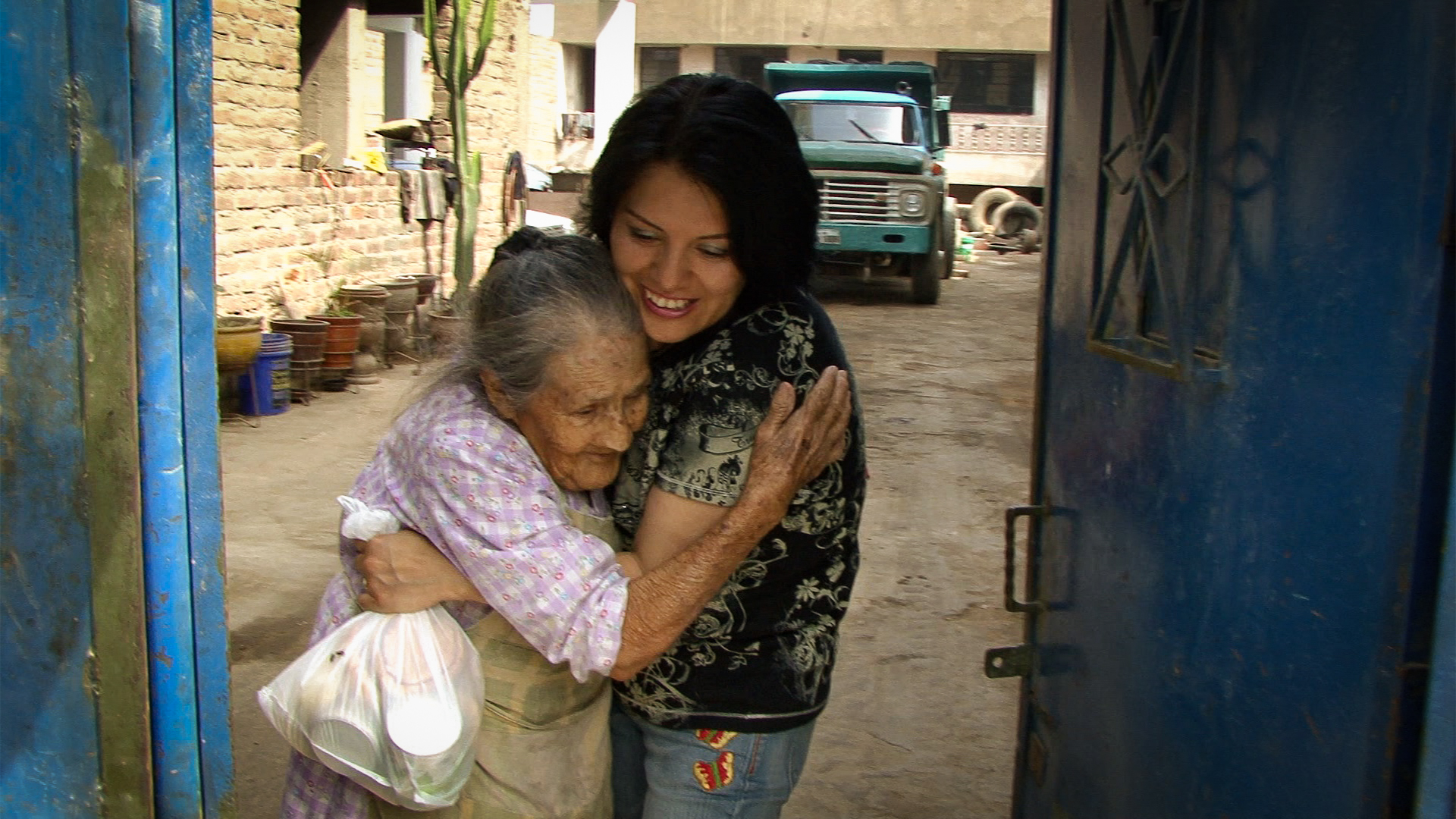 A young woman is warmly hugging an elderly woman who is holding a plastic bag. They are standing in a doorway with a truck visible in the background.
