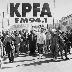 Black and white photo of a large group of people marching down a street, holding a banner that reads "KPFA FM 94.1.