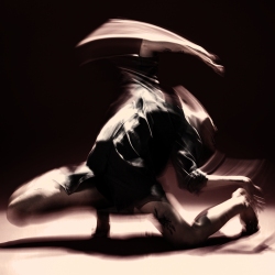A blurred image of a person in dynamic motion, with limbs extended in a dramatic dance pose against a dark background.