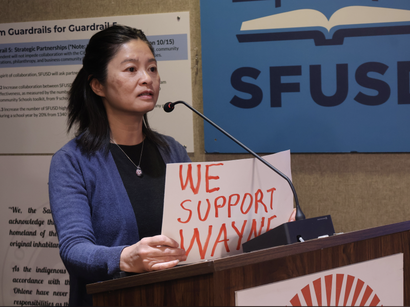 A woman speaks at a podium holding a sign that reads "We Support Wayne," with SFUSD signage in the background. Maria Su stands nearby, showing her support for the cause.