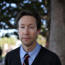 A person in a blue shirt and striped tie stands outdoors in front of a tree, looking at the camera.