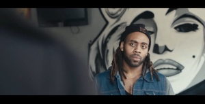 A person with long hair and a cap stands indoors, a mural of a face on the wall behind them.