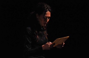 Person in a dark setting reading from a piece of paper, illuminated by a soft light.