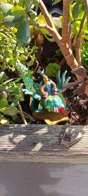 A small colorful statue of a person riding a dragon is placed among green plants in a wooden planter.