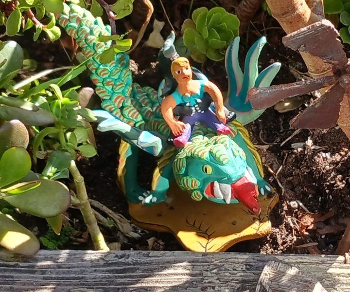 A small colorful statue of a person riding a dragon is placed among green plants in a wooden planter.