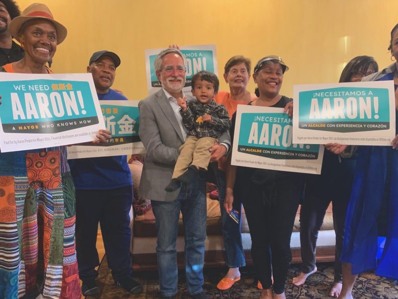 A group of people holding campaign signs that read "We Need Aaron! A Mayor Who Knows How" and "Necesitamos A Aaron! Un Alcalde Con Experiencia Y Corazon," smiling indoors.