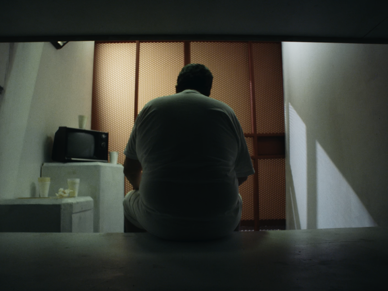 A person in a white outfit sits on a bed facing a barred window in a dimly lit room with a TV and other items on a table beside them.