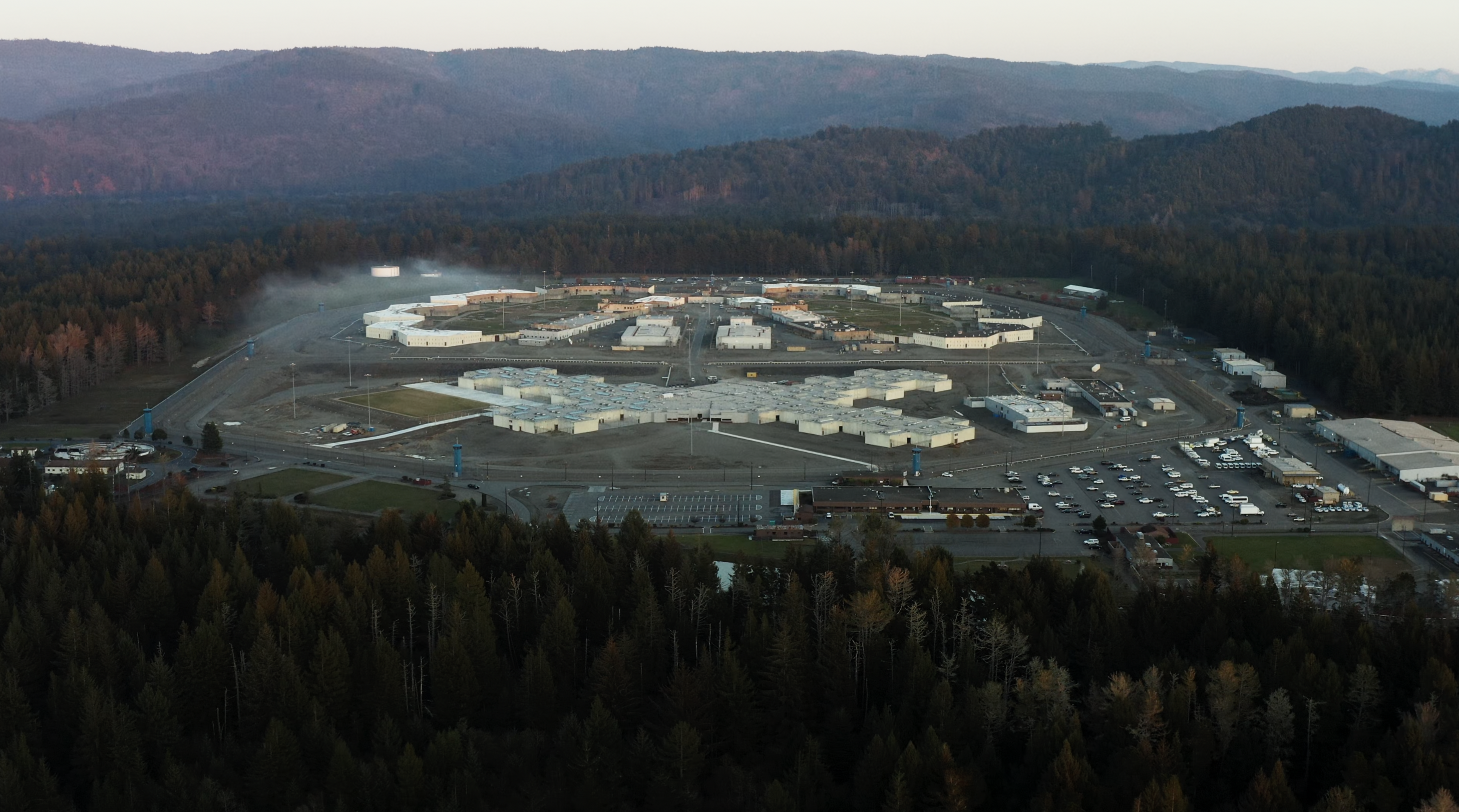 Aerial view of a large correctional facility surrounded by forested hills, with several buildings, parking lots, and roadways visible.