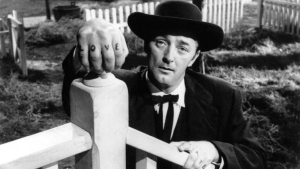 A man in a suit and hat stands by a fence, with the word "LOVE" tattooed on his fingers.