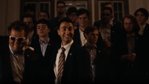 A group of people in suits and formal attire stand together indoors, with one person in the center smiling broadly.