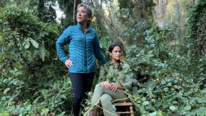 Two women in a forested area: one is standing and wearing a blue jacket, the other is seated on a wooden chair wearing green clothing, and surrounded by greenery.