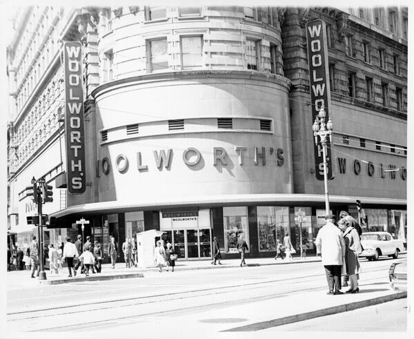 Black and white photo of a Woolworth's store on a busy city street corner with people walking and crossing.