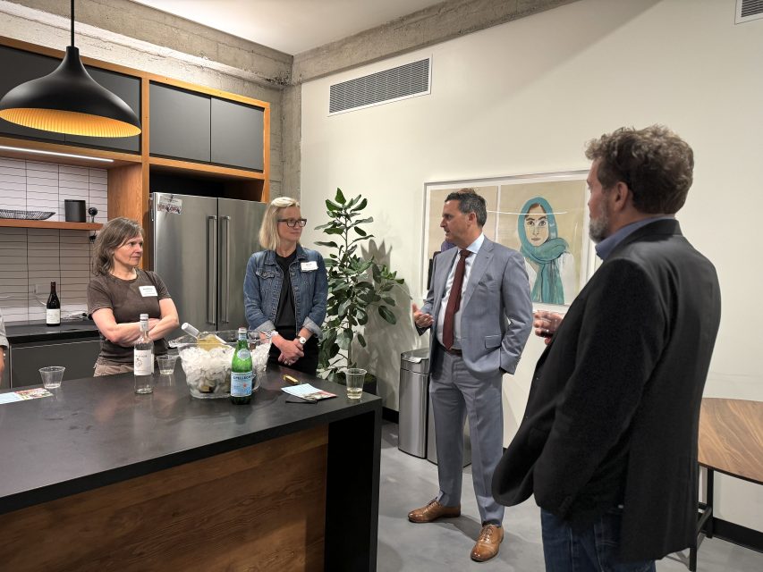 Four people in business attire are conversing in a modern kitchen area with a plant and a framed picture on the wall.