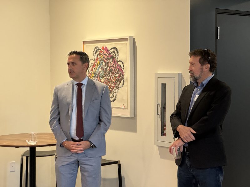 Two men stand in a room with one in a suit and the other in a blazer. There's a table, a chair, and a colorful abstract artwork on the wall.