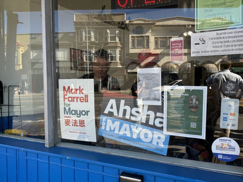 Storefront window displaying mayoral campaign posters, various notices, and QR codes in a busy street setting.