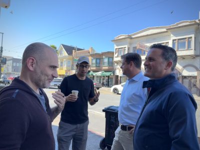Four men are talking and laughing while standing on a street with shops in the background. One man is holding a cup.