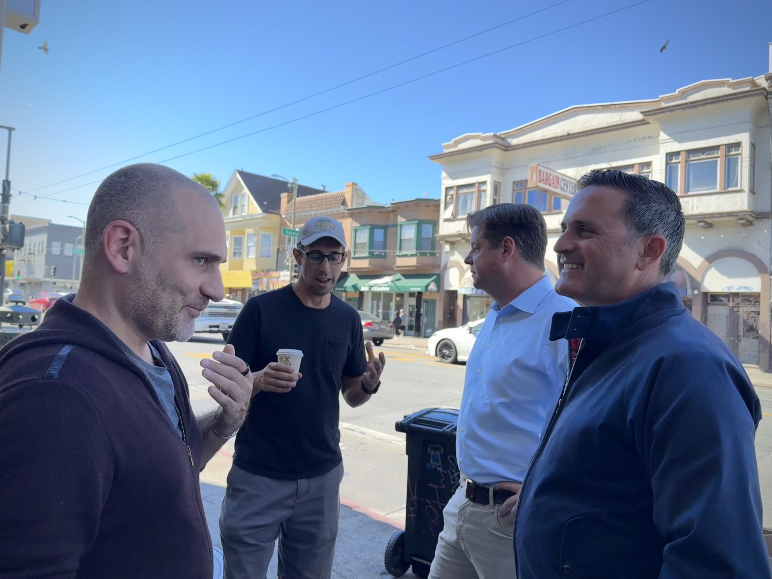 Four men are talking and laughing while standing on a street with shops in the background. One man is holding a cup.