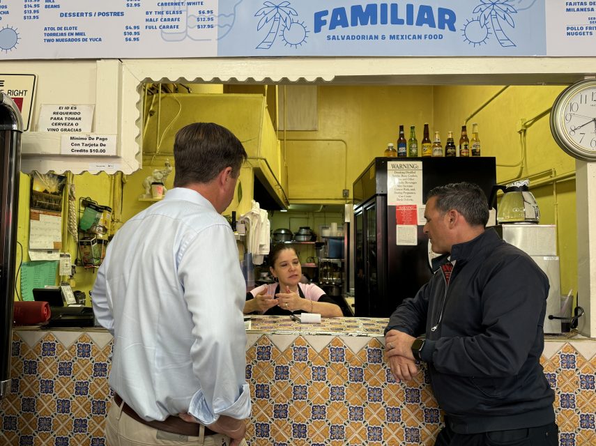 Two men in conversation with a woman at the counter of a Salvadoran and Mexican food establishment. Bottles and menu visible in the background.