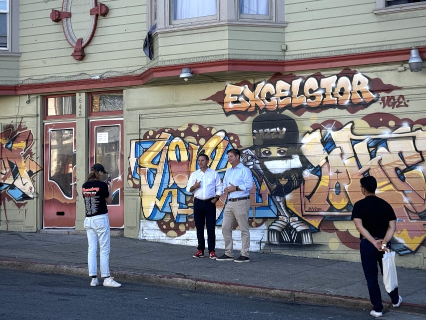Four people stand near a building with vibrant graffiti, including the word "Excelsior.