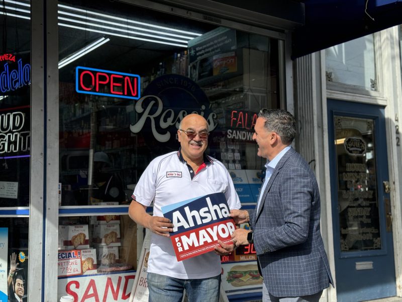 Two men stand outside a deli, smiling. One man holds a "Ahsha for Mayor" sign. A neon "Open" sign is visible above them.