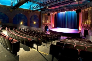 A theater with rows of red and black seats facing a stage with a blue curtain. The ceiling is painted blue with stage lights positioned above. The walls have decorative arches.