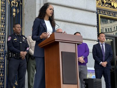 A woman speaks at a podium outside a building, with four men standing behind her, including two uniformed officers.