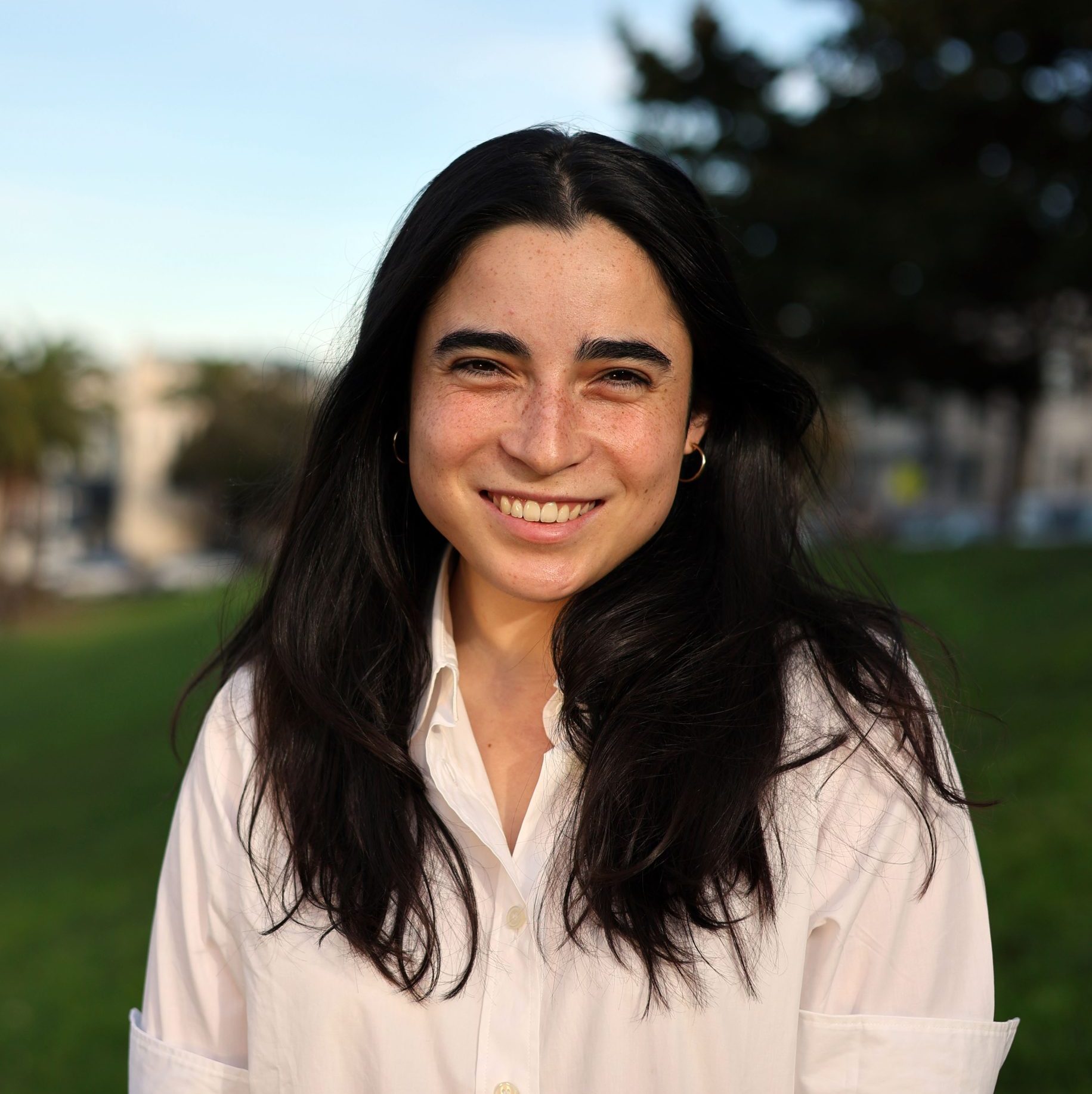 A person with long dark hair, wearing a white button-up shirt, stands outdoors on grass with trees and blurred buildings in the background, smiling at the camera.