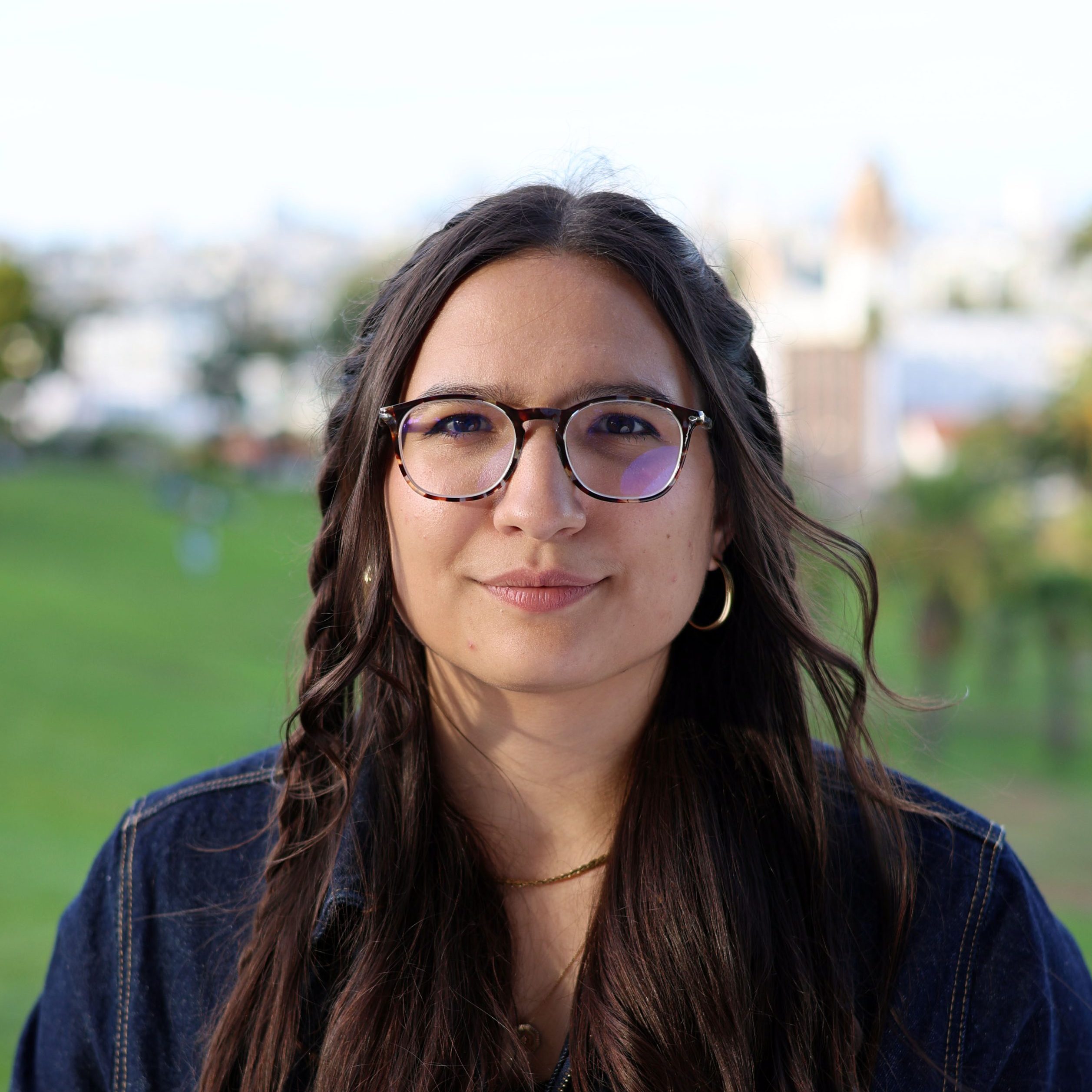 A woman with long brown hair, glasses, and a denim jacket stands outdoors with a blurred grassy area and buildings in the background.