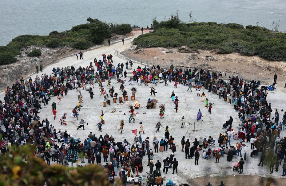 Aerial view of a large group of people gathered in a circle near the water, with some in colorful attire performing in the center. Dense greenery can be seen in the background.