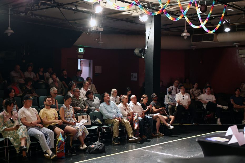A diverse group of people seated attentively in a dimly lit theater with colorful decorations hanging from the ceiling. Some hold notebooks and bags.